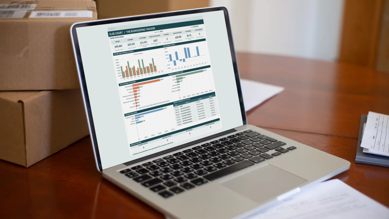 A laptop on a wooden desk beside shipping boxes, showing a small business bookkeeping dashboard with income and expense charts.